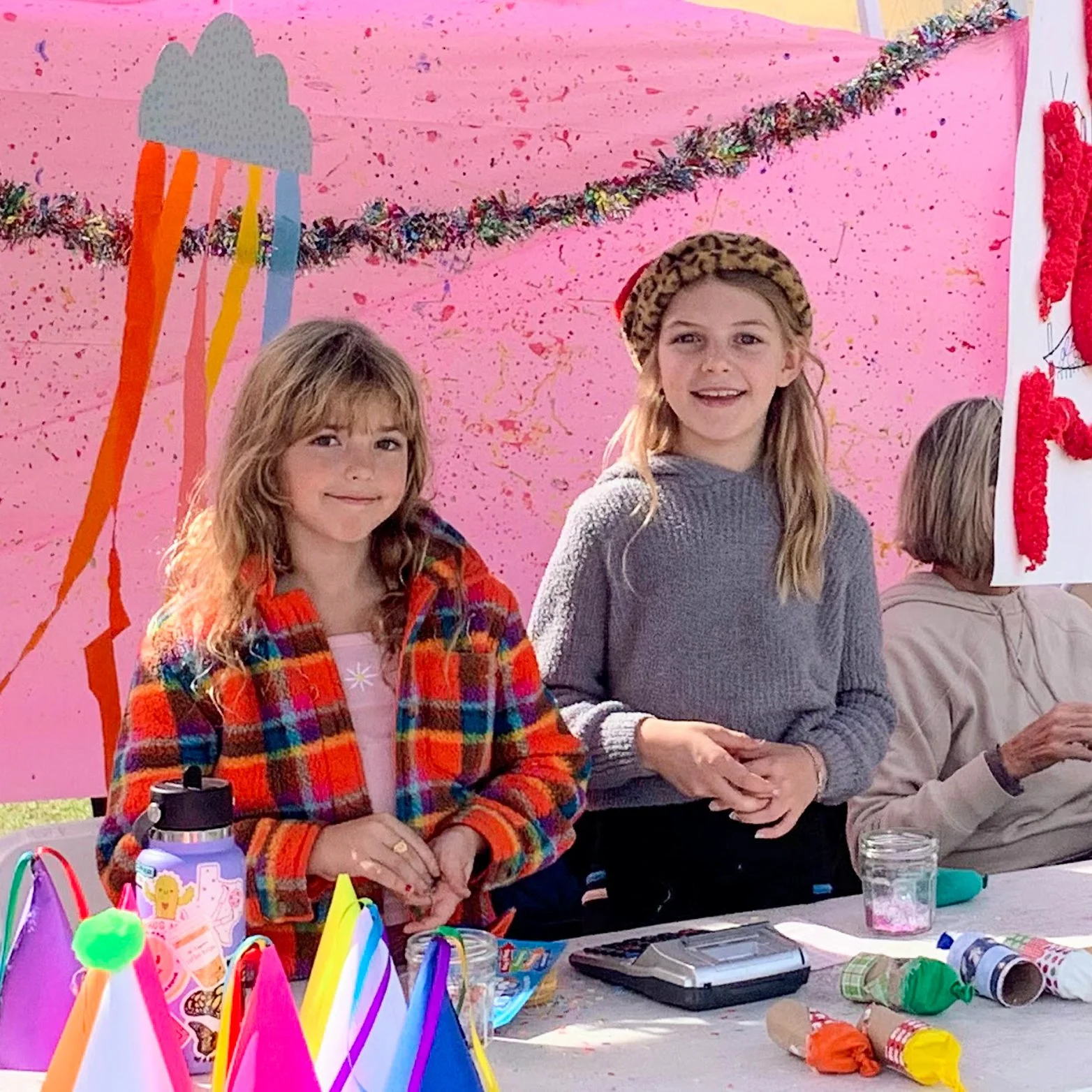 Two young girls at a birthday party, one with wavy hair wearing a colorful plaid jacket, and the other with straight hair wearing a gray sweater and leopard print headband, standing in front of a pink backdrop with a rain cloud decoration. The table in front of them has party hats, a water bottle, and party favors.