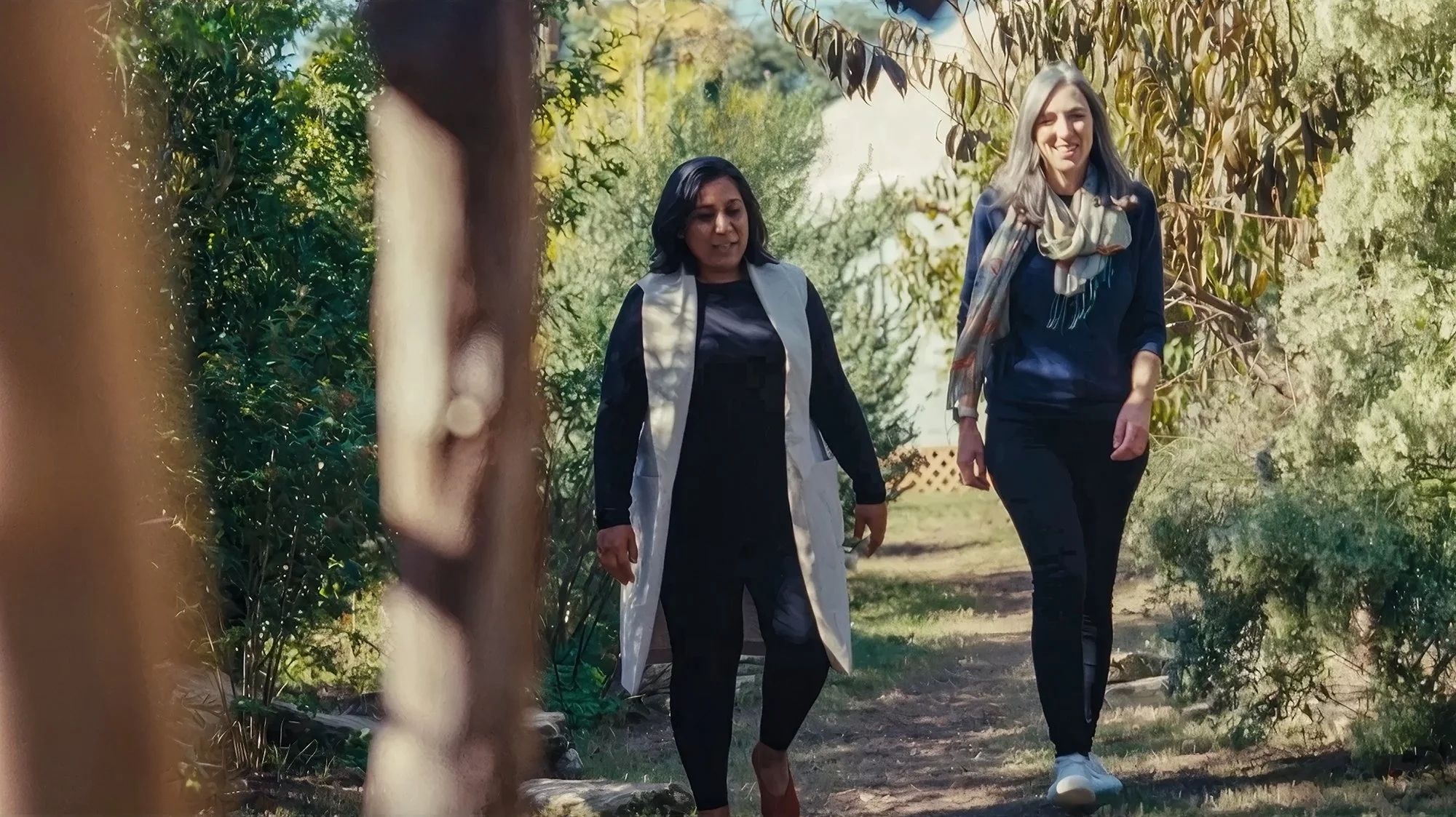 Two women walking along a wooded garden path, surrounded by green trees and bushes, enjoying a sunny day.