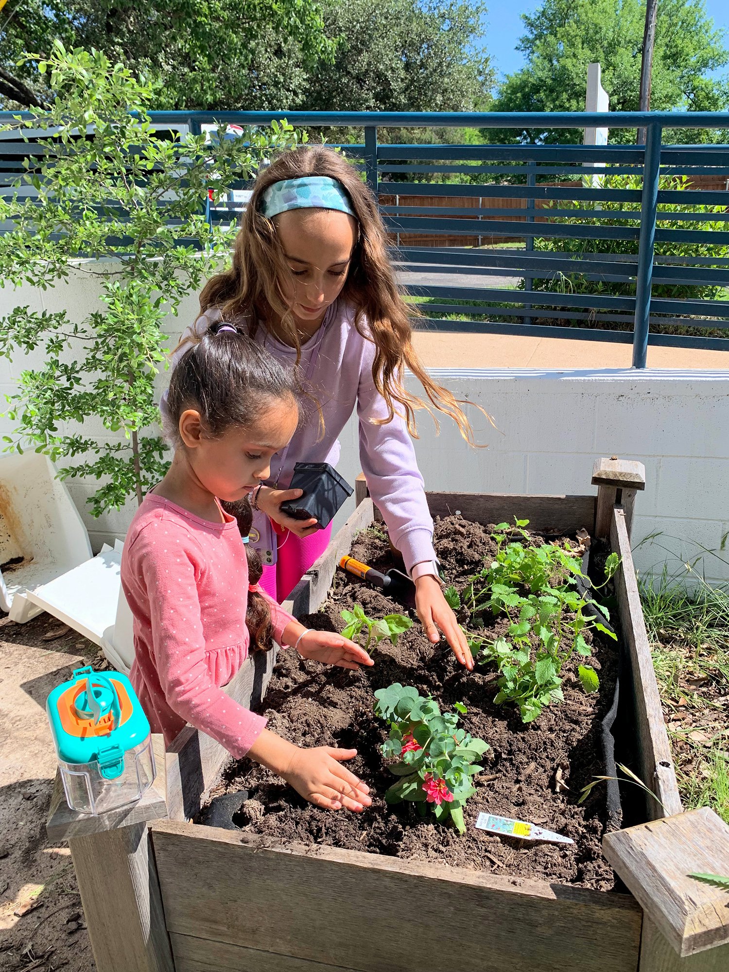 A woman and a young girl planting flowers and vegetables in a raised garden bed outdoors on a sunny day.