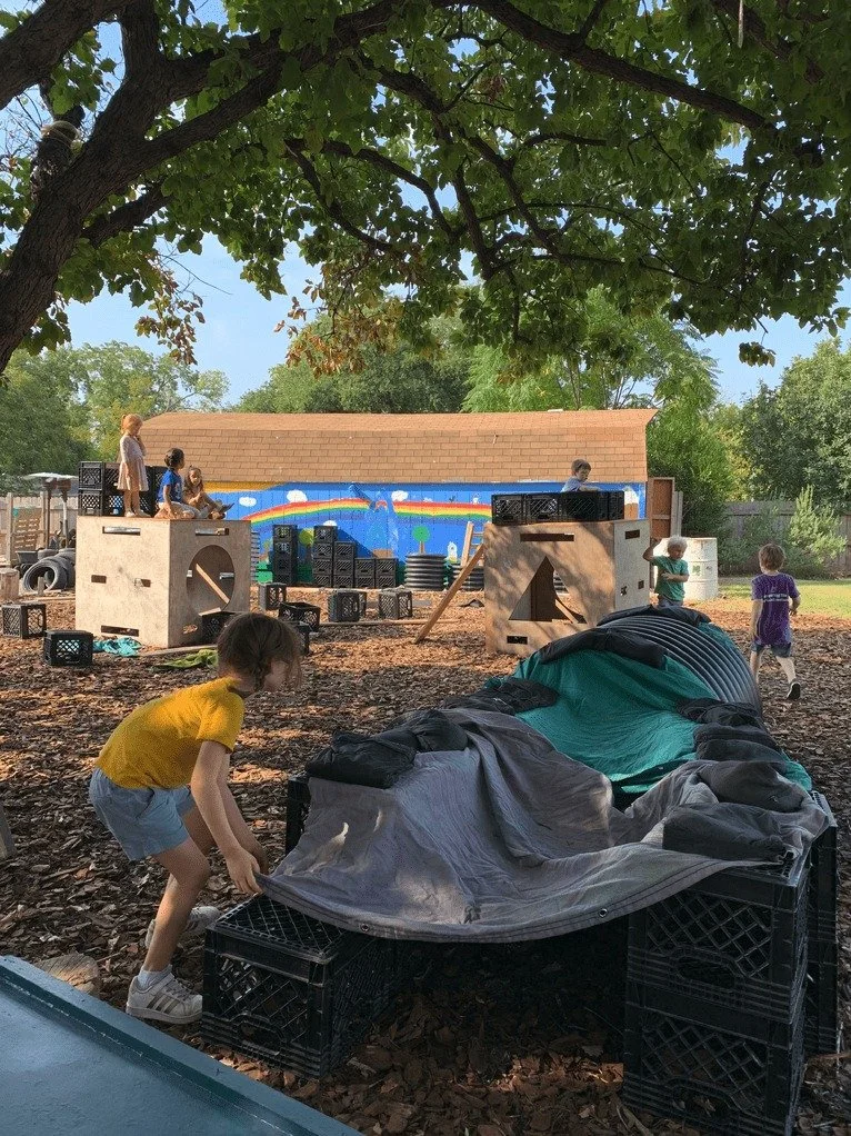 Children playing outdoors on a playground under a large tree with green leaves, with a painted mural of a rainbow and clouds in the background, and a tent set up on the ground.