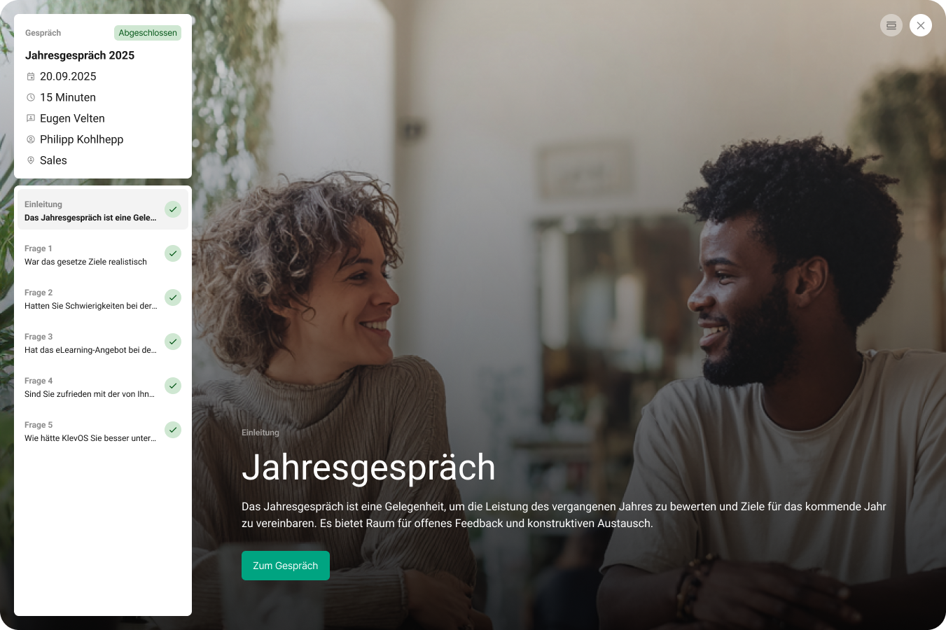 Zwei Menschen, eine Frau mit lockigem Haar und ein Mann mit krausem Haar, lächeln während eines Gesprächs an einem Tisch in einem gut beleuchteten Raum. Im Hintergrund sind Pflanzen und ein Fenster sichtbar.