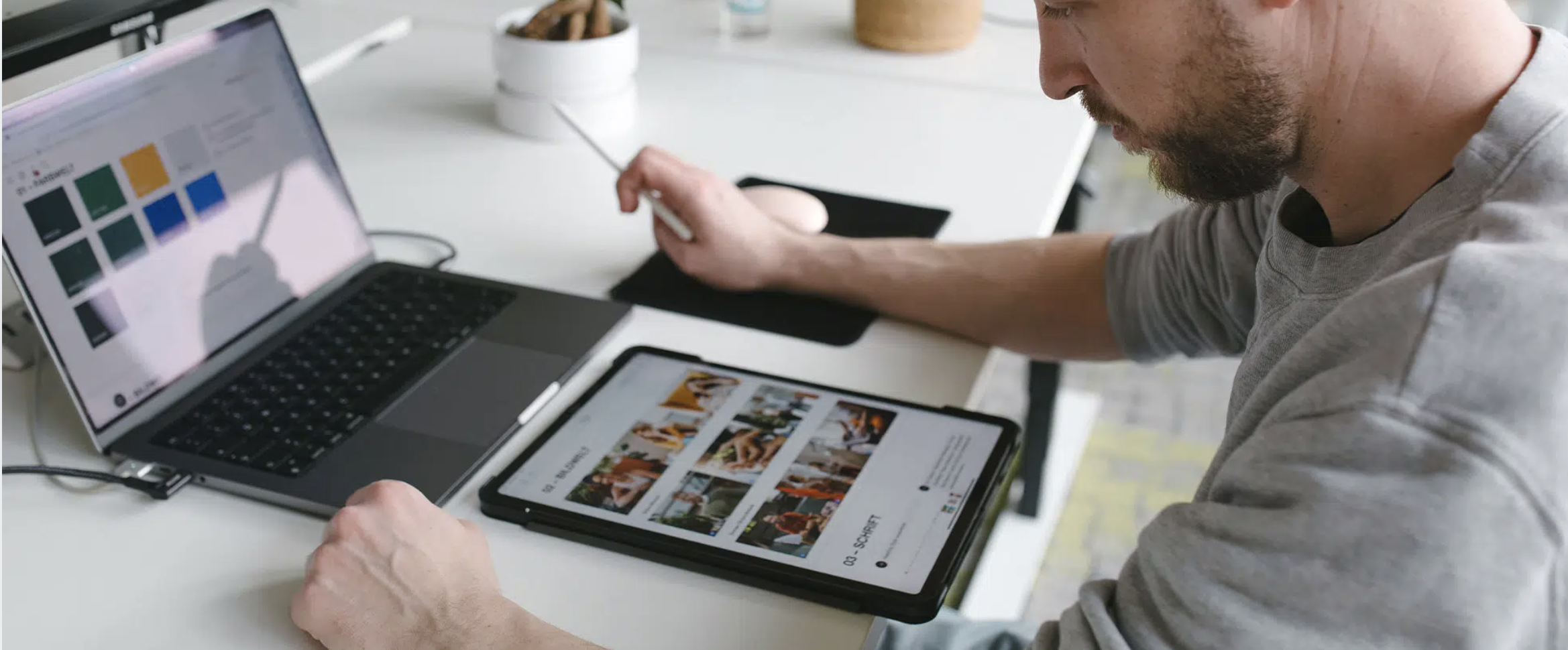 Ein Mann arbeitet an einem Laptop und Tablet auf einem Schreibtisch, während er mit einem Stift auf dem Tablet zeichnet, mit Pflanzen im Hintergrund.