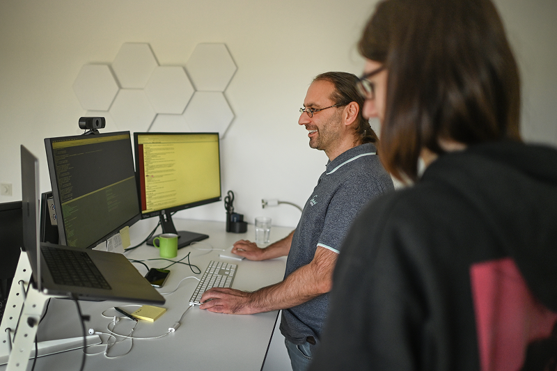 Zwei Männer arbeiten in einem Büro an Computern mit zwei Monitoren, die Programmiercode anzeigen, während sie sprechen und lachen.
