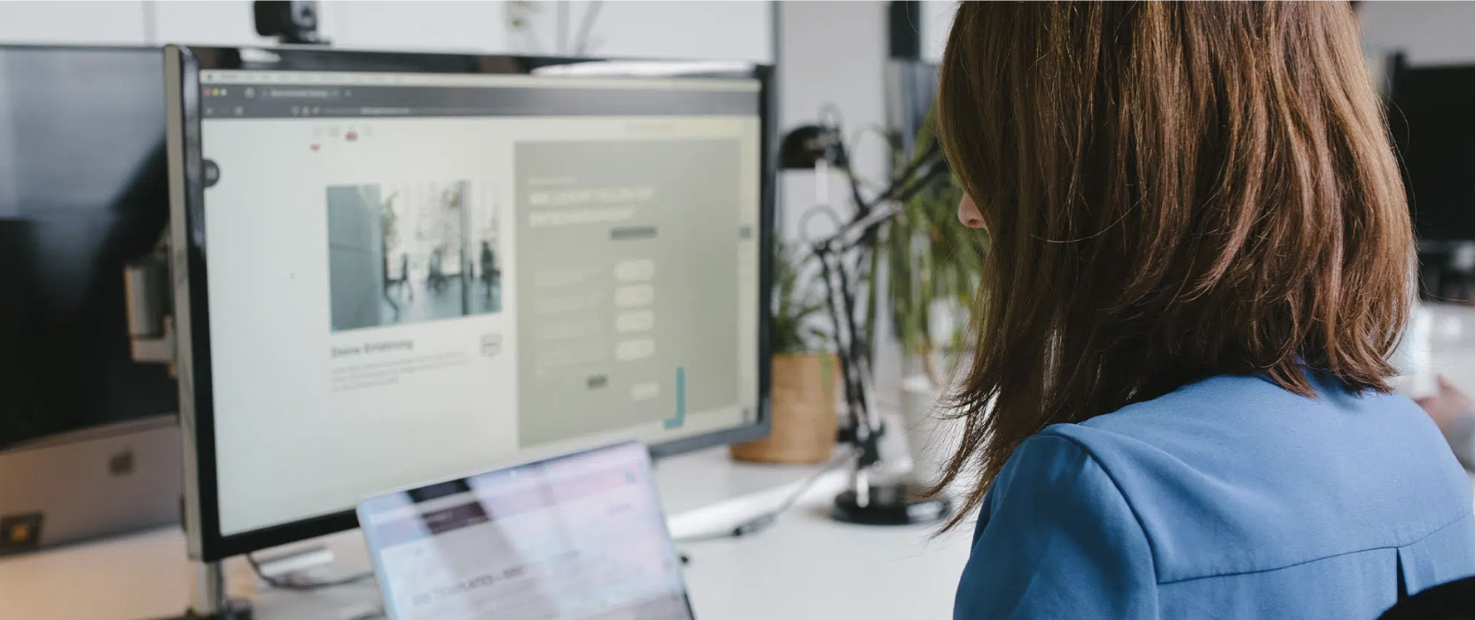 Frau mit braunen, gewellten Haaren sitzt an einem Schreibtisch, arbeitet an einem Computer und blickt auf den Bildschirm.