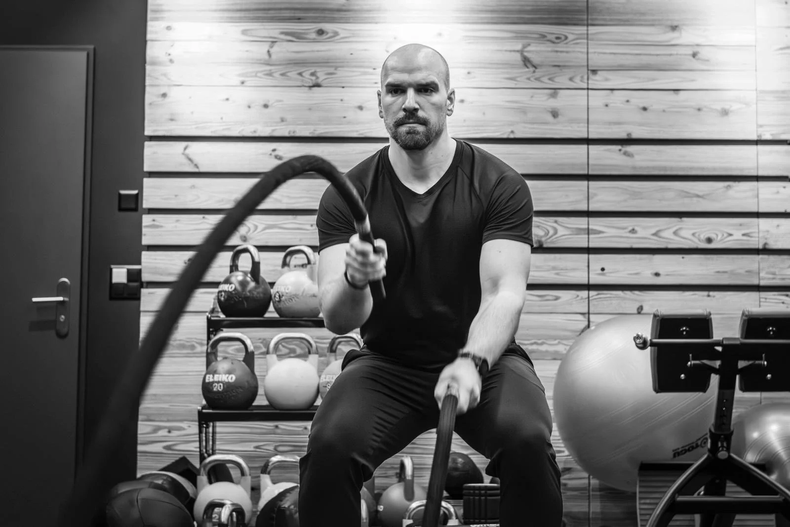 Ein Mann trainiert mit einem Seil in einem Fitnessstudio, mit Hanteln, Kettlebells und Gymball im Hintergrund.