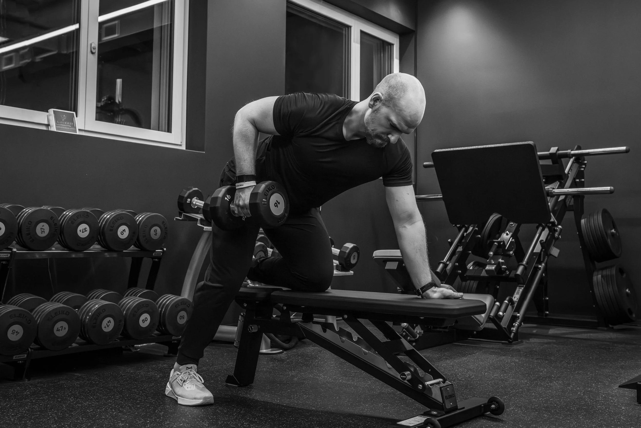 Ein Mann trainiert im Fitnessstudio mit einem Kurzhantel, während er auf einer Bank in einer schwarzen Kleidung steht.