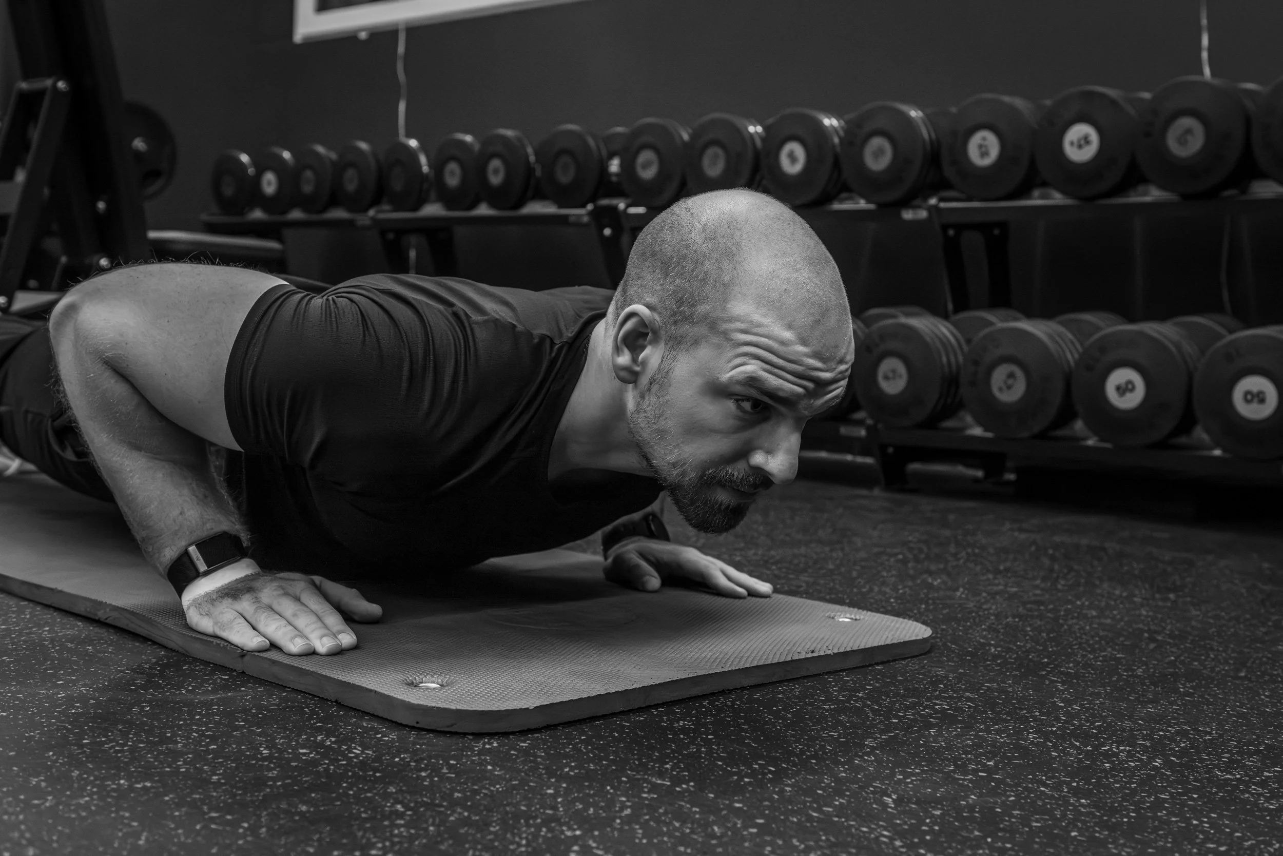 Ein Mann macht Liegestütze auf einer Yogamatte im Fitnessstudio, im Hintergrund sind Hanteln auf einem Regal. Das Bild ist schwarz-weiß.