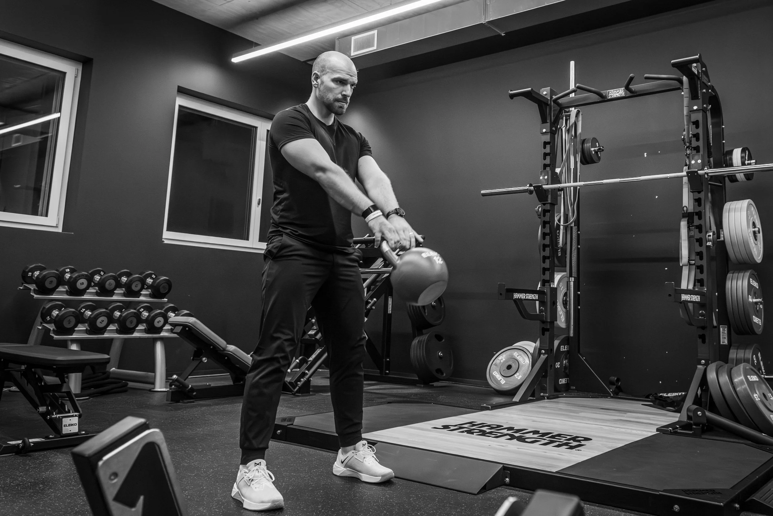 Ein Mann hebt mit beiden Händen eine Kettlebell in einem Fitnessstudio. Im Hintergrund sind Hanteln, Gewichte und ein Squat-Rack zu sehen.
