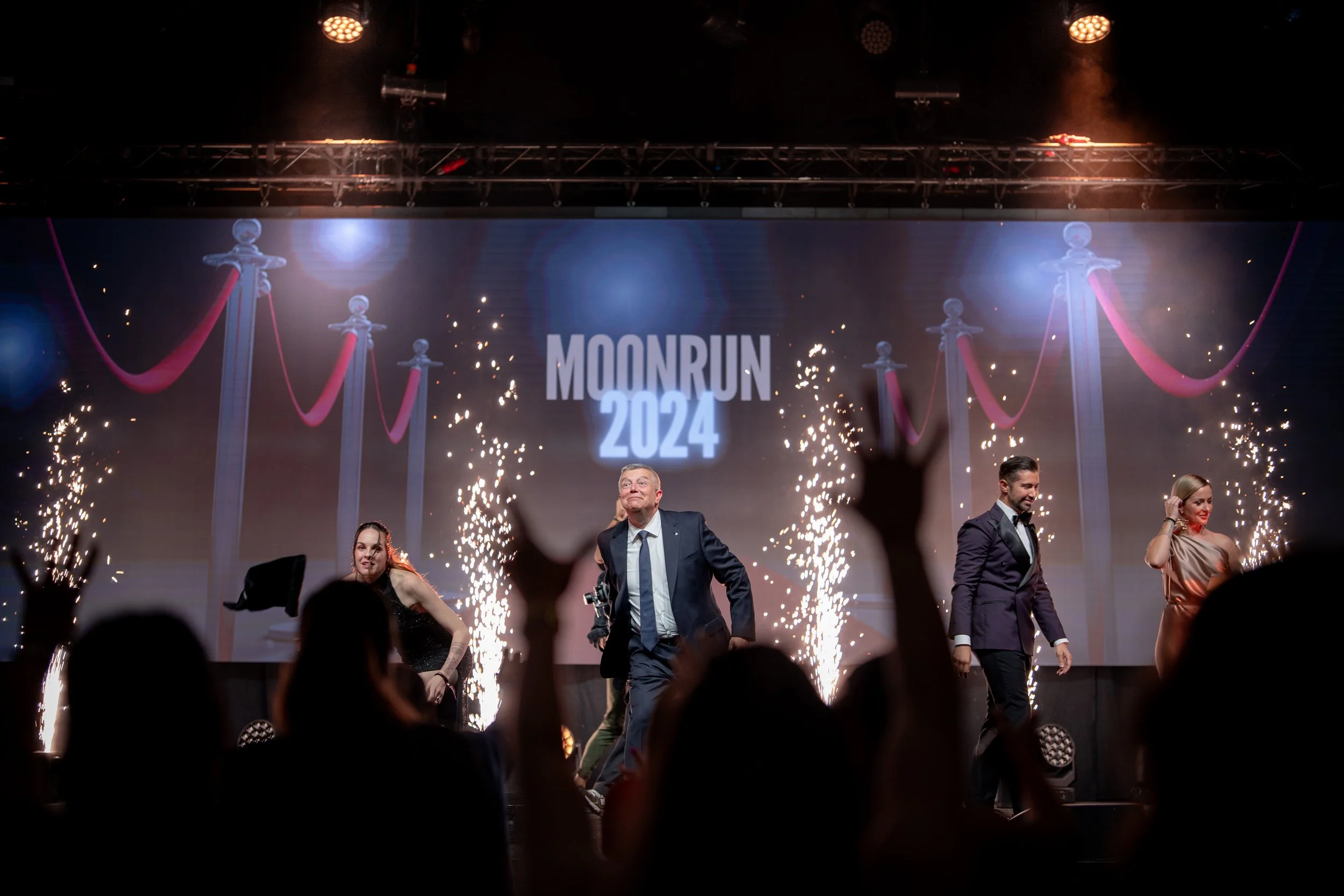 Menschen auf einer Bühne beim Moonrun 2024, umgeben von Feuerwerken, mit einem großen Bildschirm im Hintergrund, der den Namen und das Jahr zeigt.
