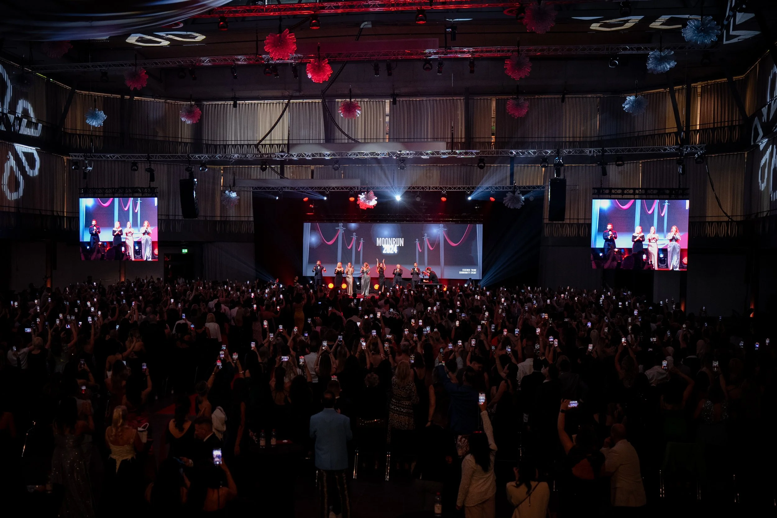 Ein großer Saal mit einer Bühne, auf der mehrere Menschen auftreten. Die Bühne ist mit roten Dekorationen geschmückt, und es gibt große Bildschirme, die die Darsteller zeigen. Das Publikum nimmt Fotos mit ihren Handys auf.