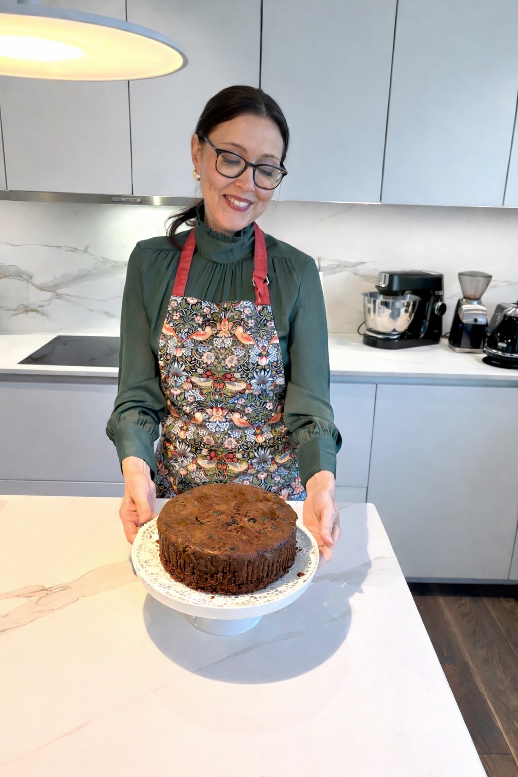 A woman in a green blouse and colorful apron holding a chocolate cake on a white cake stand in a modern kitchen.