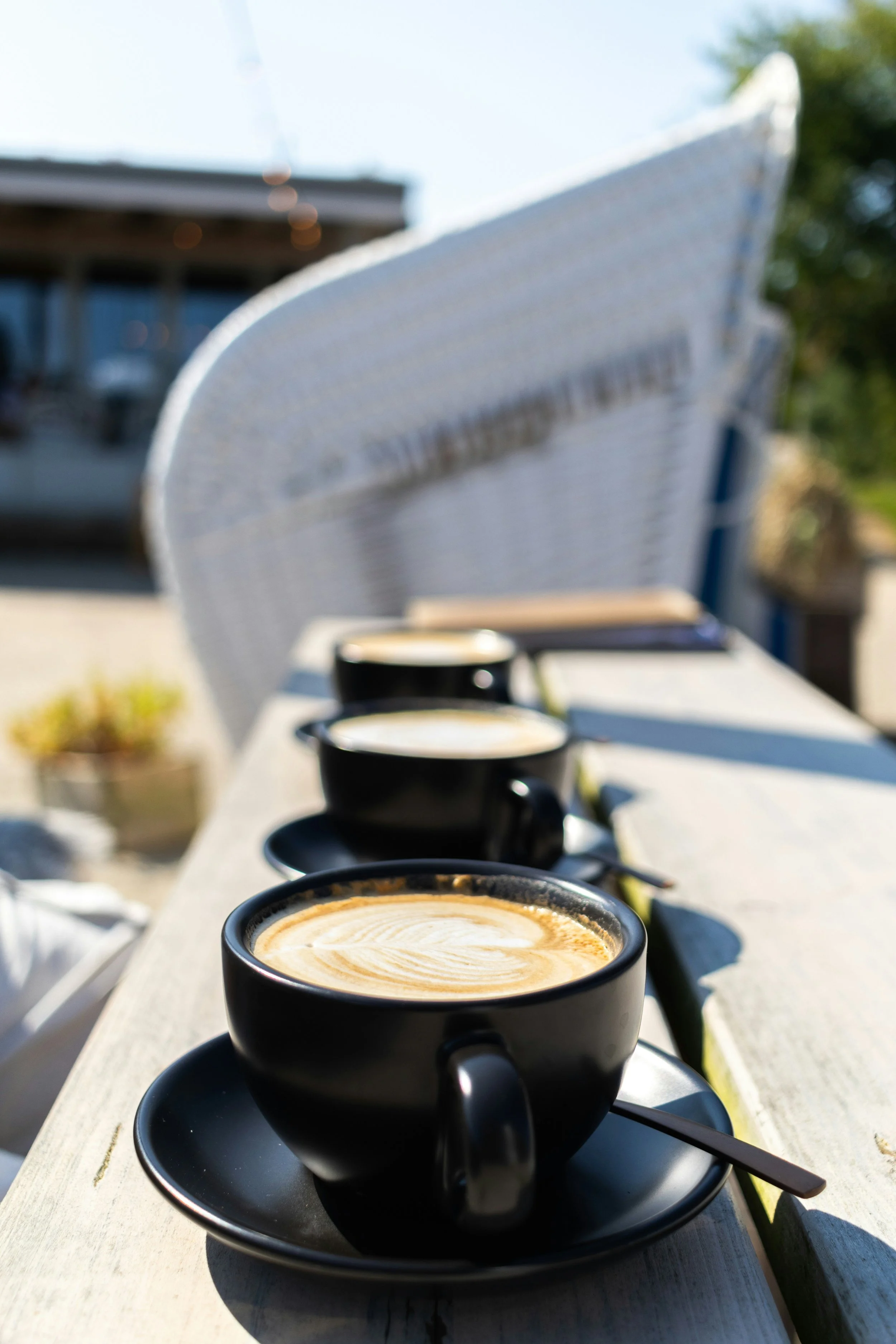 Three black coffee cups with latte art on a white outdoor table, with chairs in the background and a sunny outdoor setting.