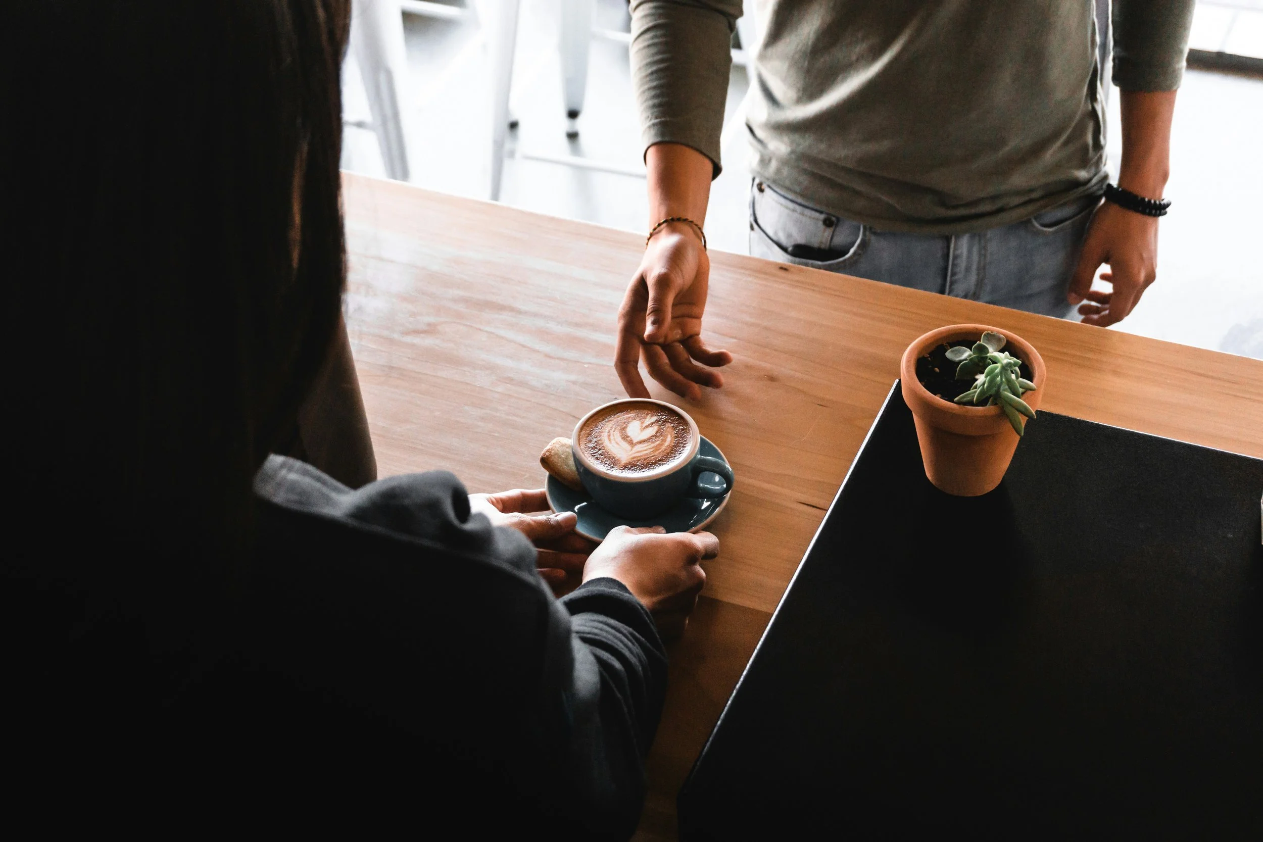 Person sitting at a wooden table with a cup of coffee, a sweet treat, and a potted plant, while another person stands nearby reaching for the cup.