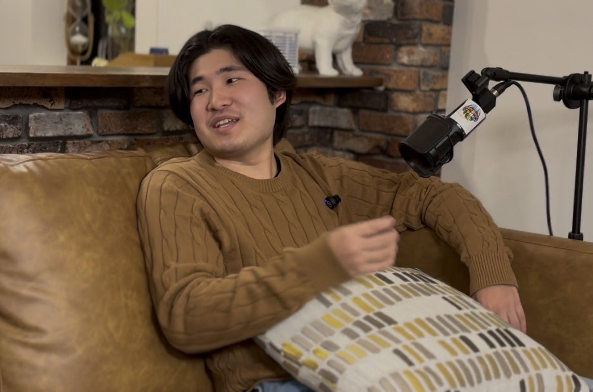 A young man with dark hair is sitting on a brown leather couch, talking, with a pillow on his lap. Behind him, there is a brick wall with a shelf that has decorative items, including a white ceramic cat.