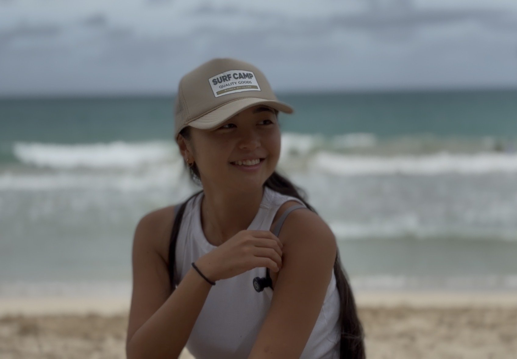 Smiling woman on beach wearing a beige cap labeled 'Surf Camp' and a sleeveless white top with sunglasses hanging from her shirt.