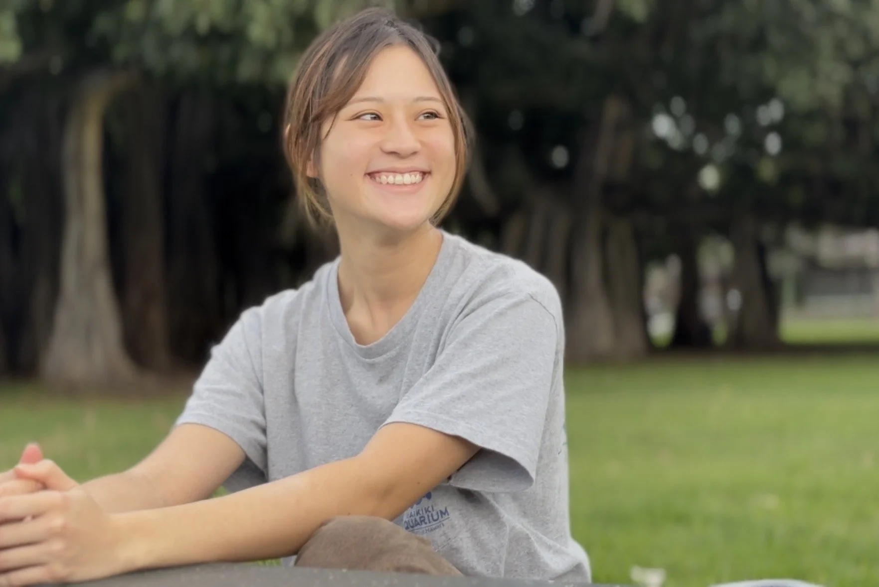 A young woman with short brown hair smiling while sitting outdoors on grass with large trees in the background.
