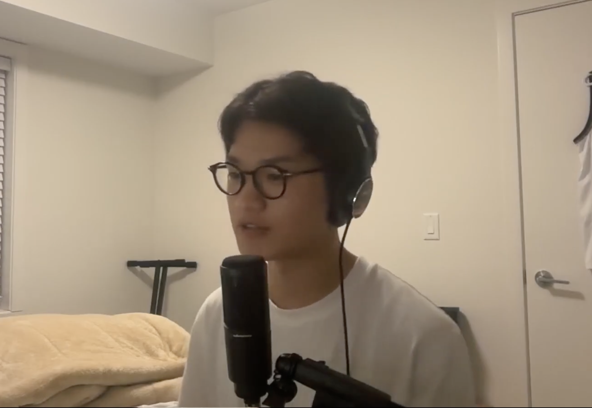 Young person with glasses and dark hair wearing headphones, speaking into a microphone in a room with a bed, a door, and an ironing board.