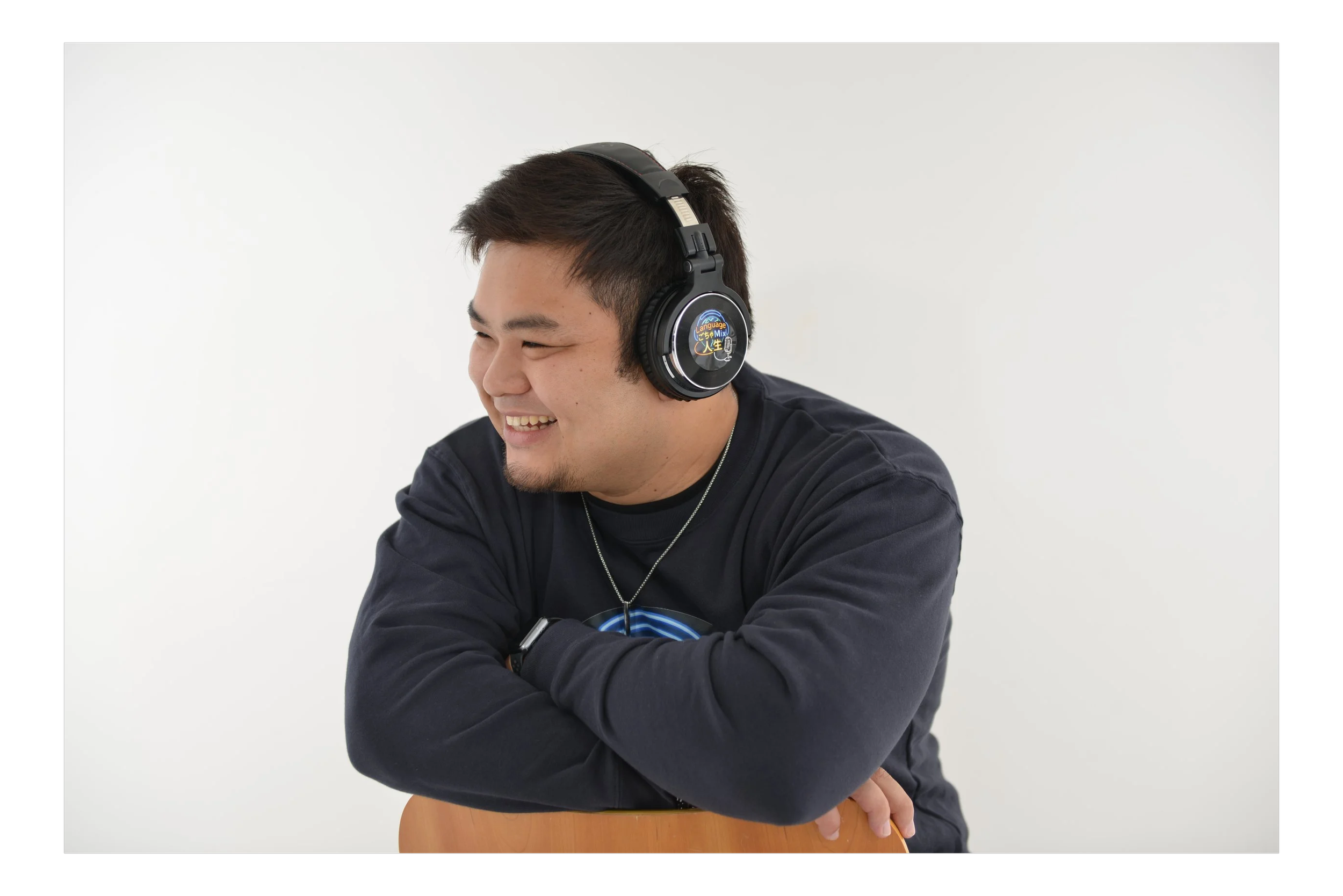 Young man smiling, wearing black headphones, and a black sweatshirt, sitting at a table against a white background.