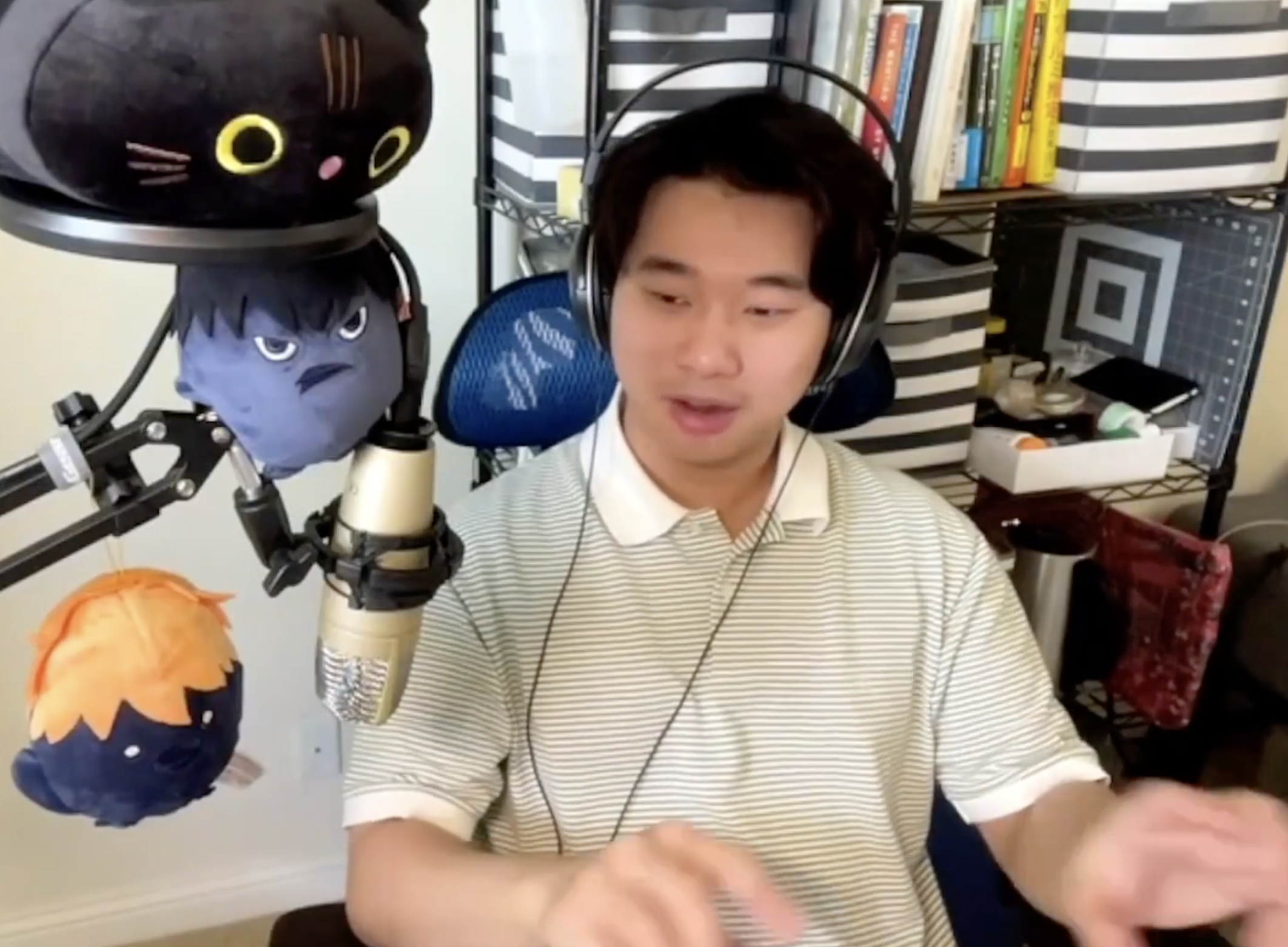 A young man wearing headphones sits at a desk with a microphone in front of him. There are plush toys with faces hanging from a microphone arm, and a bookshelf in the background filled with books and storage boxes.