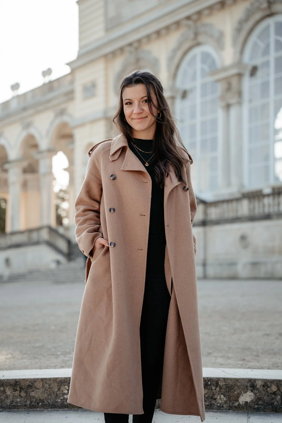 Eine junge Frau trägt einen langen beigefarbenen Mantel, steht vor einem historischen Gebäude mit großen Fenstern und Statuen im Hintergrund. Ernährungswissenschaften und Ernährungsberatung und Fitness.