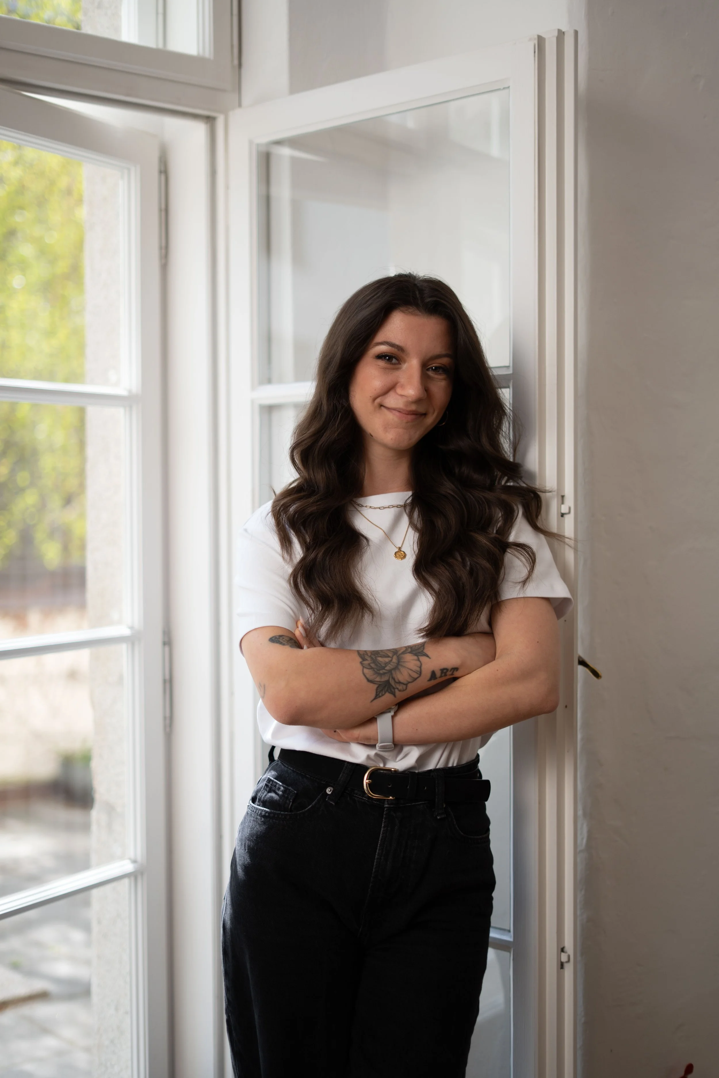 Junge und fitte Ernährungsberaterin mit dunklen, gewellten Haaren und Tattoo auf dem Arm steht lächelnd vor einem Fenster