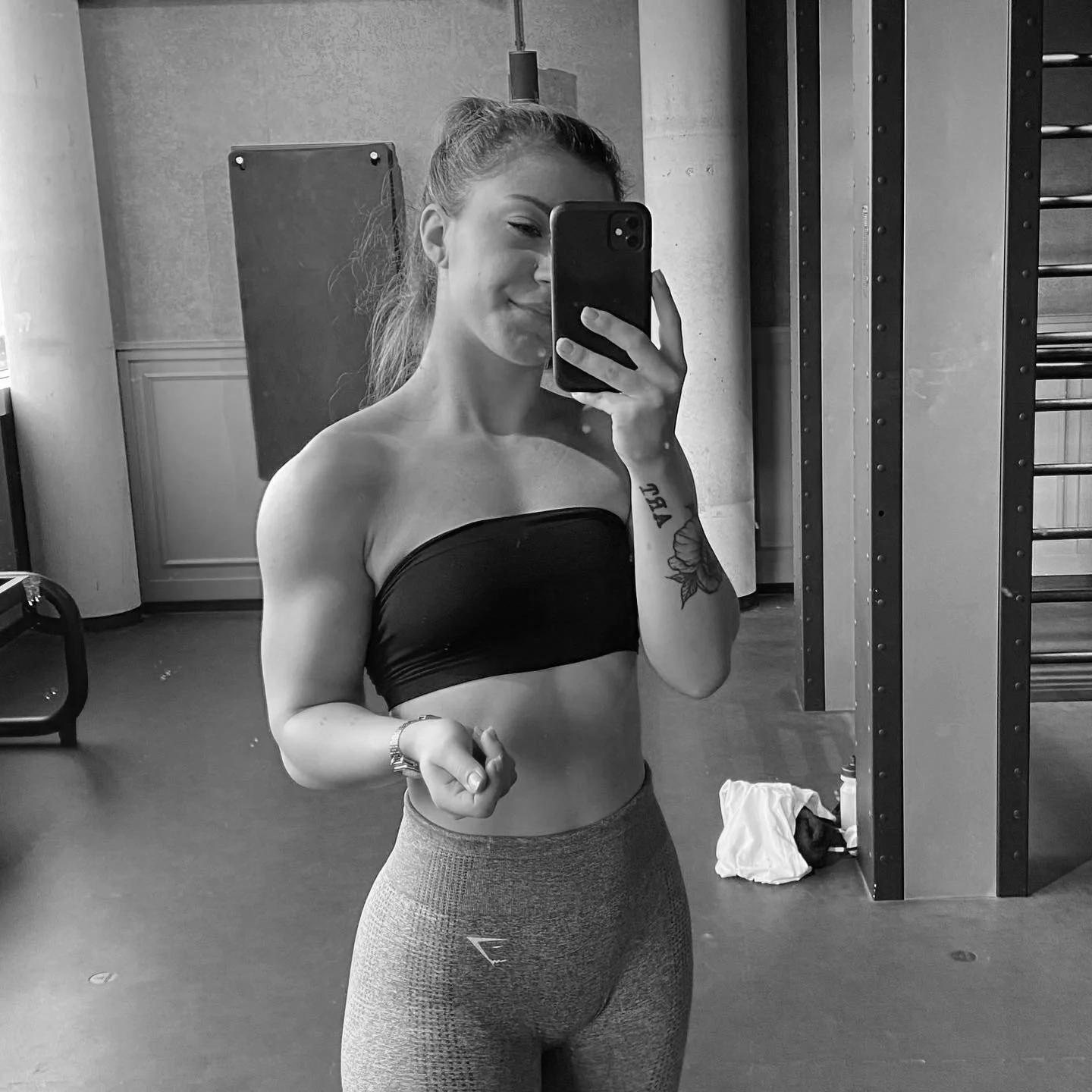 Junge Frau macht Selfie im Fitnessstudio, trägt Sportkleidung, graue Leggings und ein schwarzes Top, ohne Schuhe, schwarz-weiß Fotografie. Ernährungswissenschaften und Ernährungsberatung und Fitness.