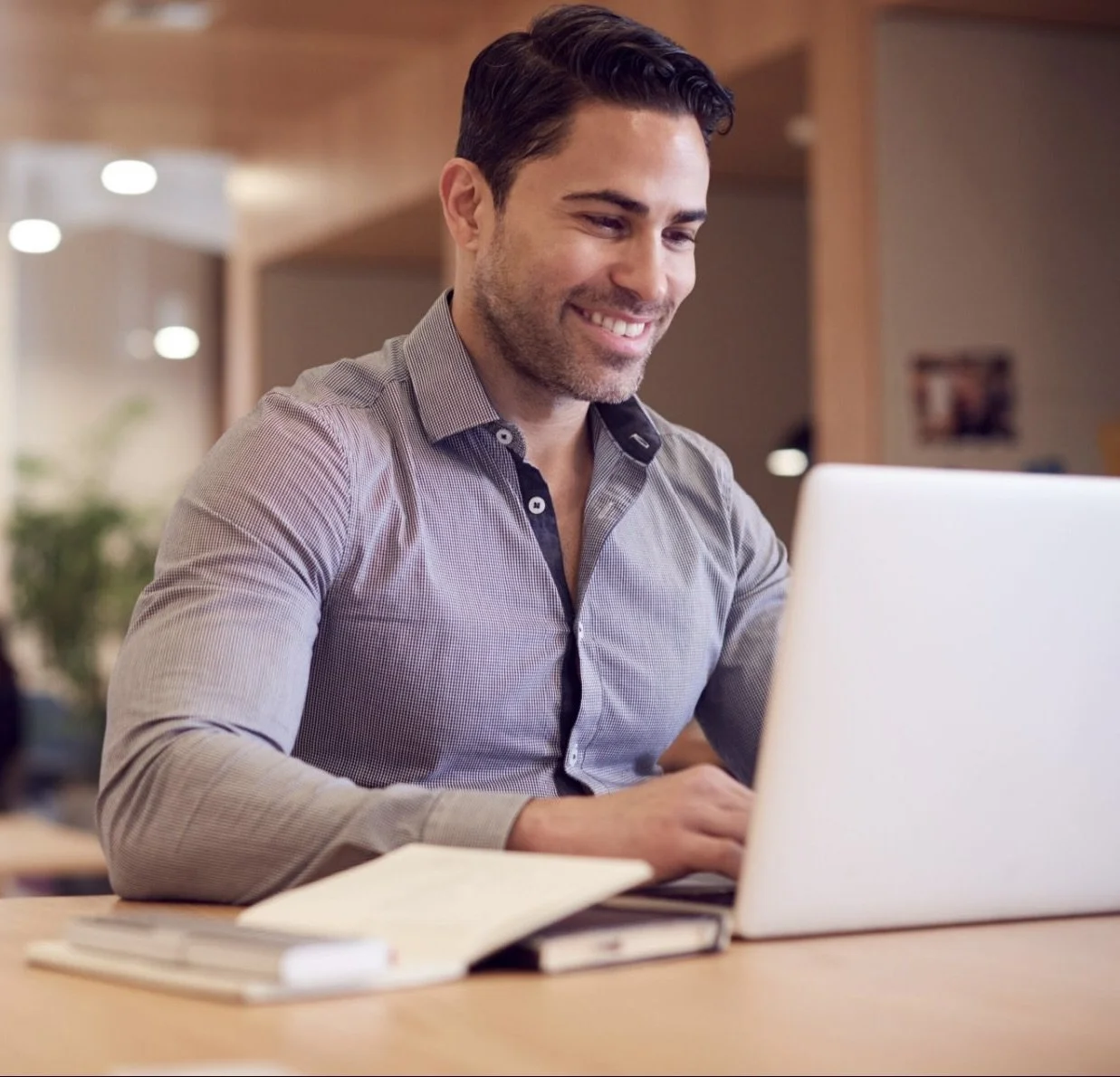 A smiling man with dark hair working on a laptop at a desk in a modern office environment.