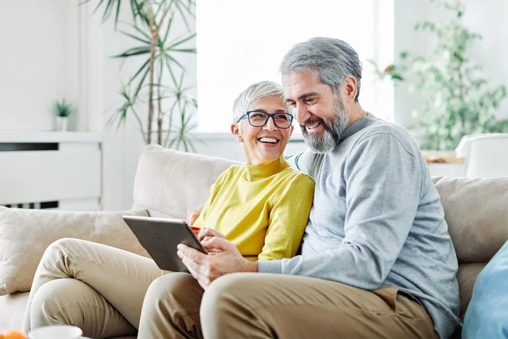 An elderly couple sitting on a beige sofa, smiling and looking at a tablet together in a bright, modern living room with large windows, green plants, and minimal decor.