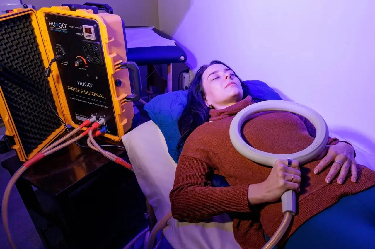 A woman lying on a bed with her eyes closed, holding a circular medical device connected to a large yellow machine beside her.