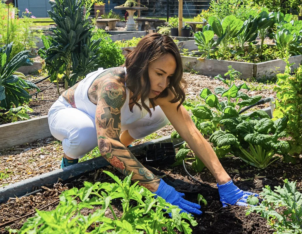 A woman with tattoos planting or tending to plants in a garden with various leafy green vegetables.