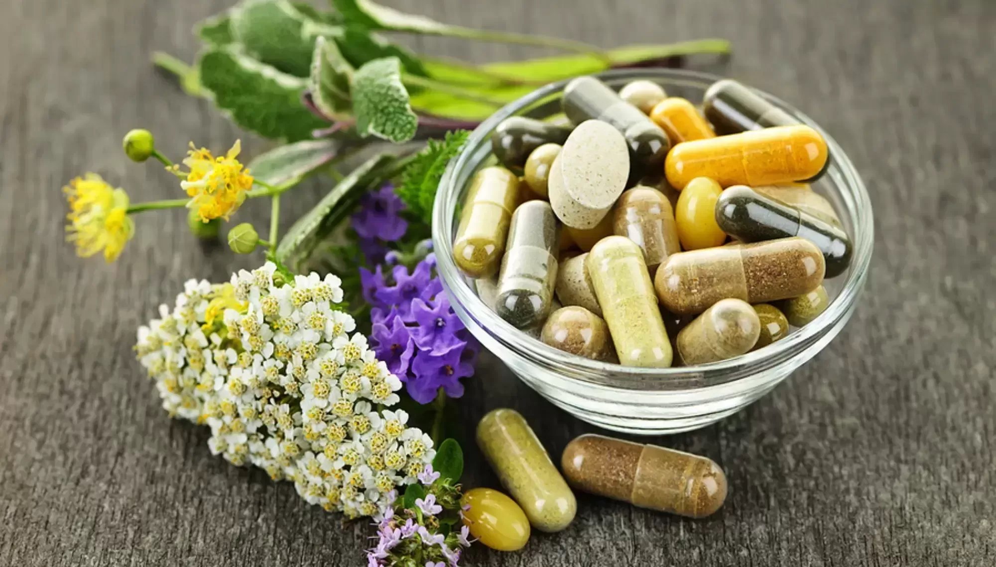 A glass bowl filled with various capsules and tablets, placed on a wooden surface with an assortment of fresh herbs and flowers beside it.