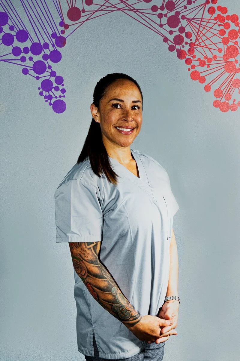 A smiling woman in a light gray medical scrub top standing in front of a wall with colorful abstract DNA helix designs in purple and red.