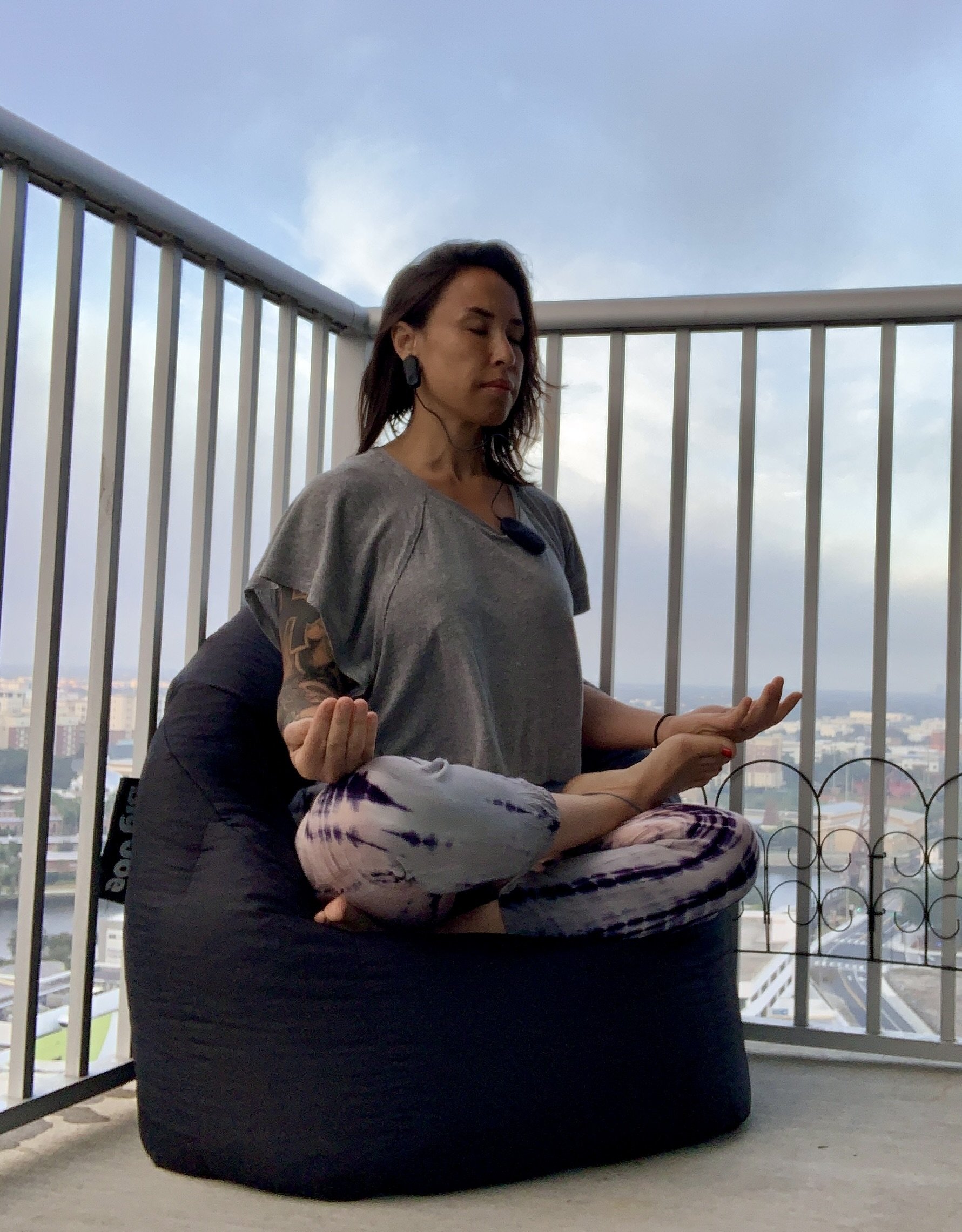 Woman meditating on a balcony with city view, sitting cross-legged on a bean bag, in a meditative pose with eyes closed.