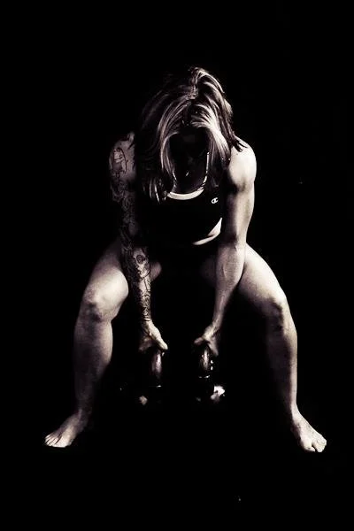 A woman with tattoos lifting kettlebells in a dark setting.