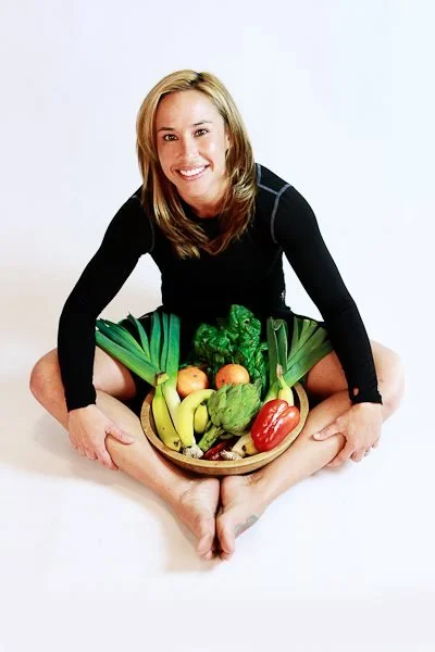 Woman sitting cross-legged on the floor holding a basket of fresh fruits and vegetables, smiling at the camera.