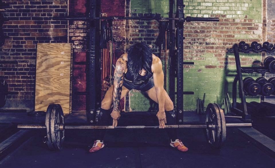 A woman with tattoos on her arms lifting a barbell in a gym with brick walls and workout equipment.