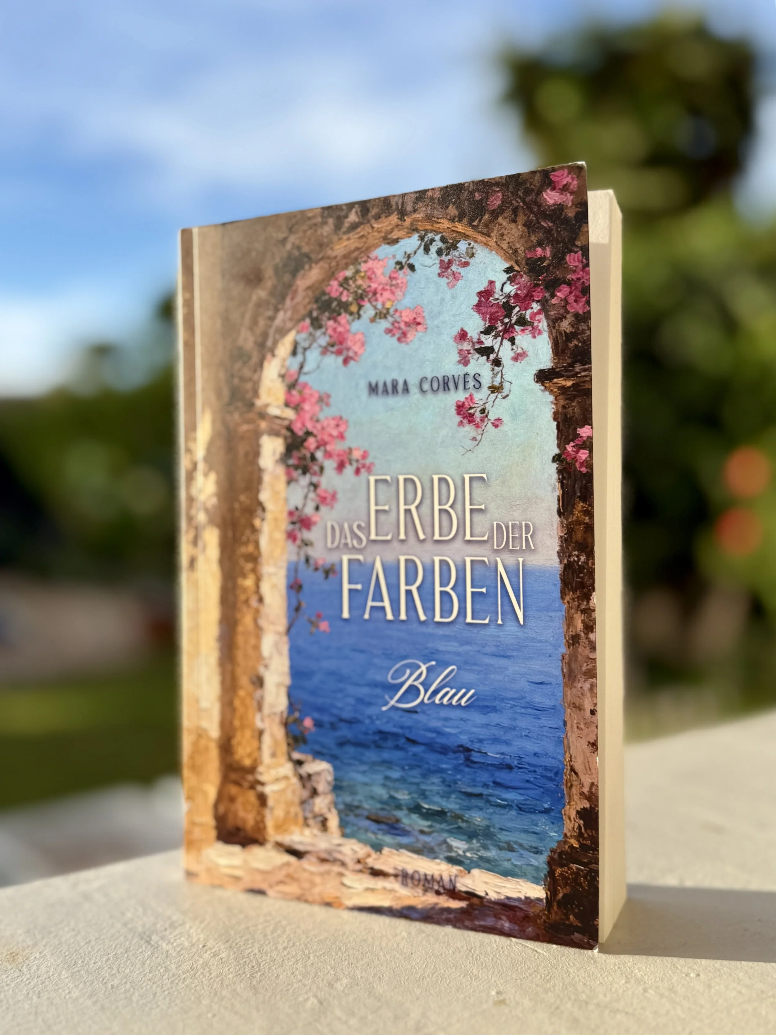 Buch mit dem Titel 'Das Erbe der Farben Blau' von Mara Corvès, das auf einer weißen Oberfläche steht, mit einem blauen Himmel und grünen Bäumen im Hintergrund.