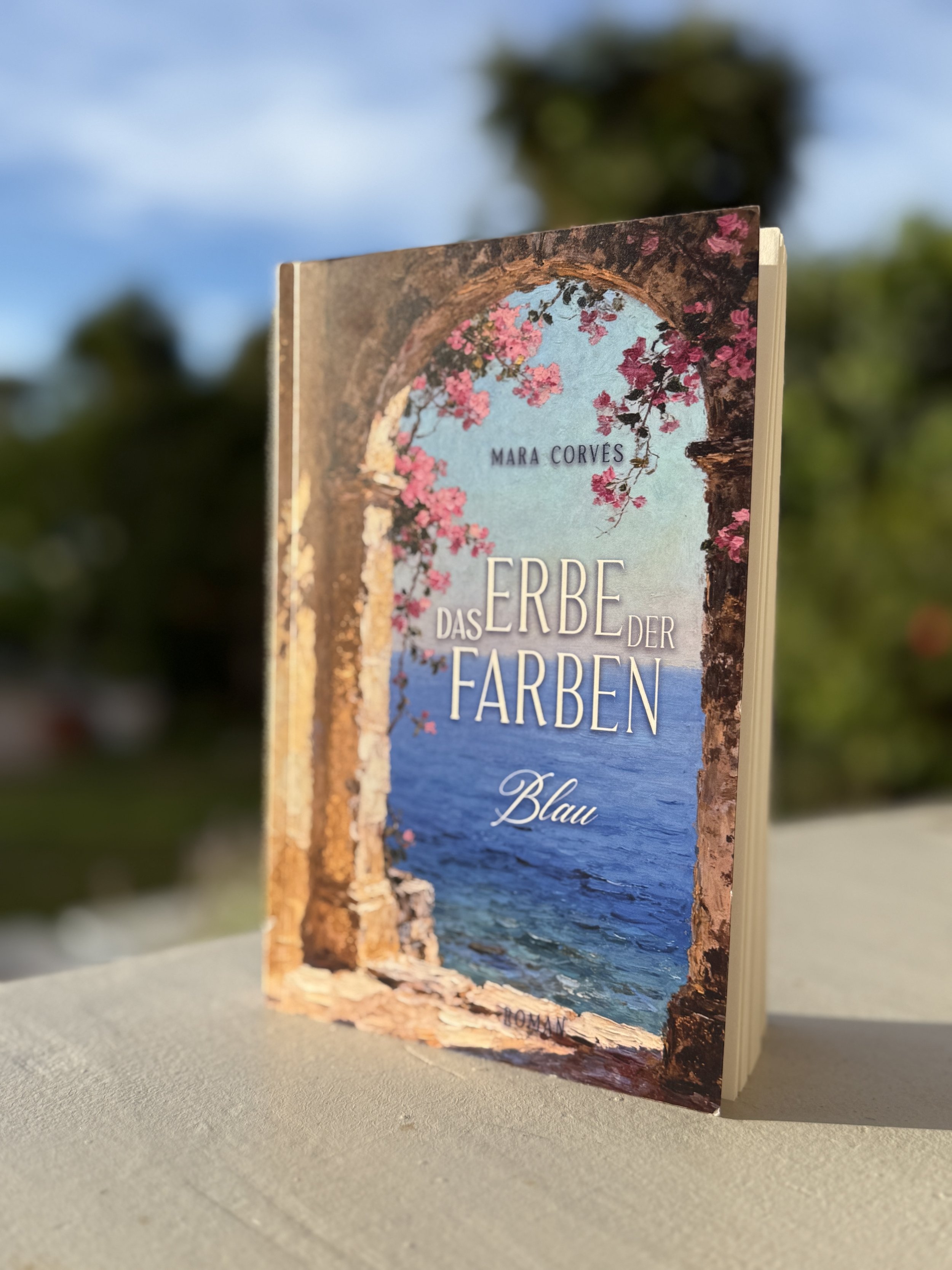 Buch mit dem Titel 'Das Erbe der Farben: Blau' von Mara Corves, zeigt ein Cover mit einem Meeresblick, einem Bogen mit blühenden pinken Blumen und einer alten Steinmauer im Sonnenschein.
