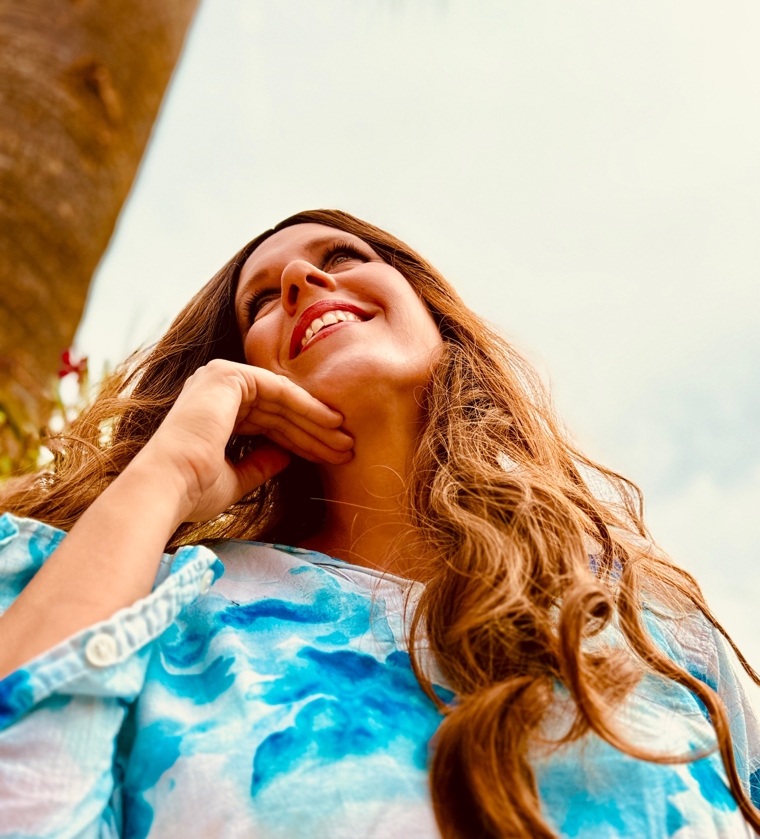 Eine lachende Frau mit langen braunen Haaren schaut nach oben, trägt ein blau-weißes Kleid, und befindet sich im Freien bei bewölktem Himmel.