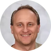 Portret van een lachende man met korte, lichte haar en een lichte huid, tegen een lichte achtergrond.