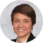 Een portret van een vrouw met kort bruin haar, glimlachend, met een witte blouse en een zwarte jas.