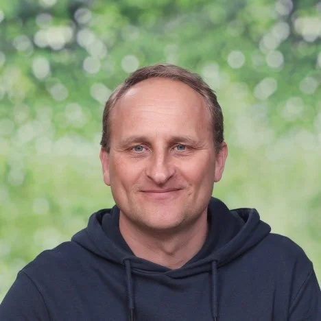 Portret van een lachende man met korte, lichte haar en een lichte huid, tegen een lichte achtergrond.