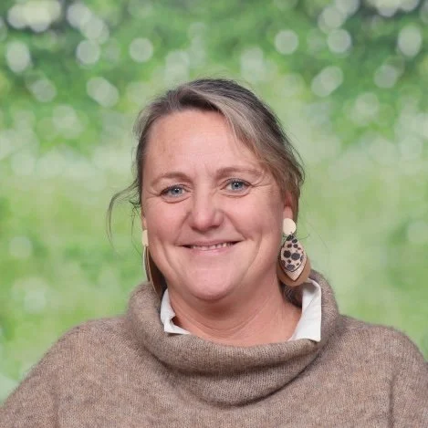 Een close-up van een lachende vrouw met kort blond haar, earrings en een roze vest.