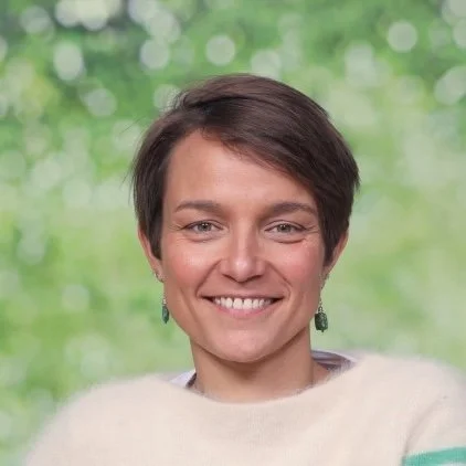 Een portret van een vrouw met kort bruin haar, glimlachend, met een witte blouse en een zwarte jas.