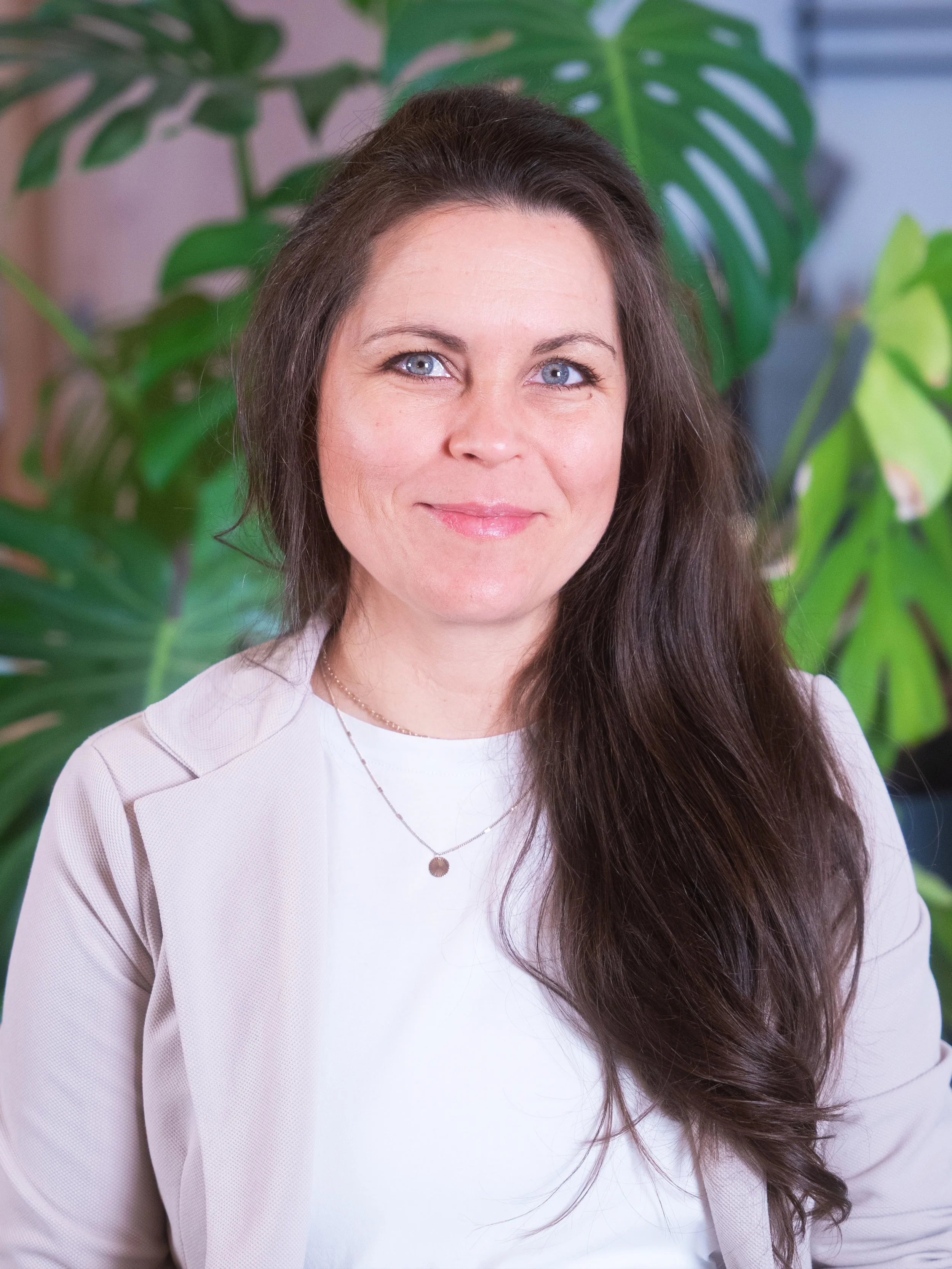 Porträt einer lächelnden Frau mit braunen, gewellten Haaren vor grünen Pflanzblättern.