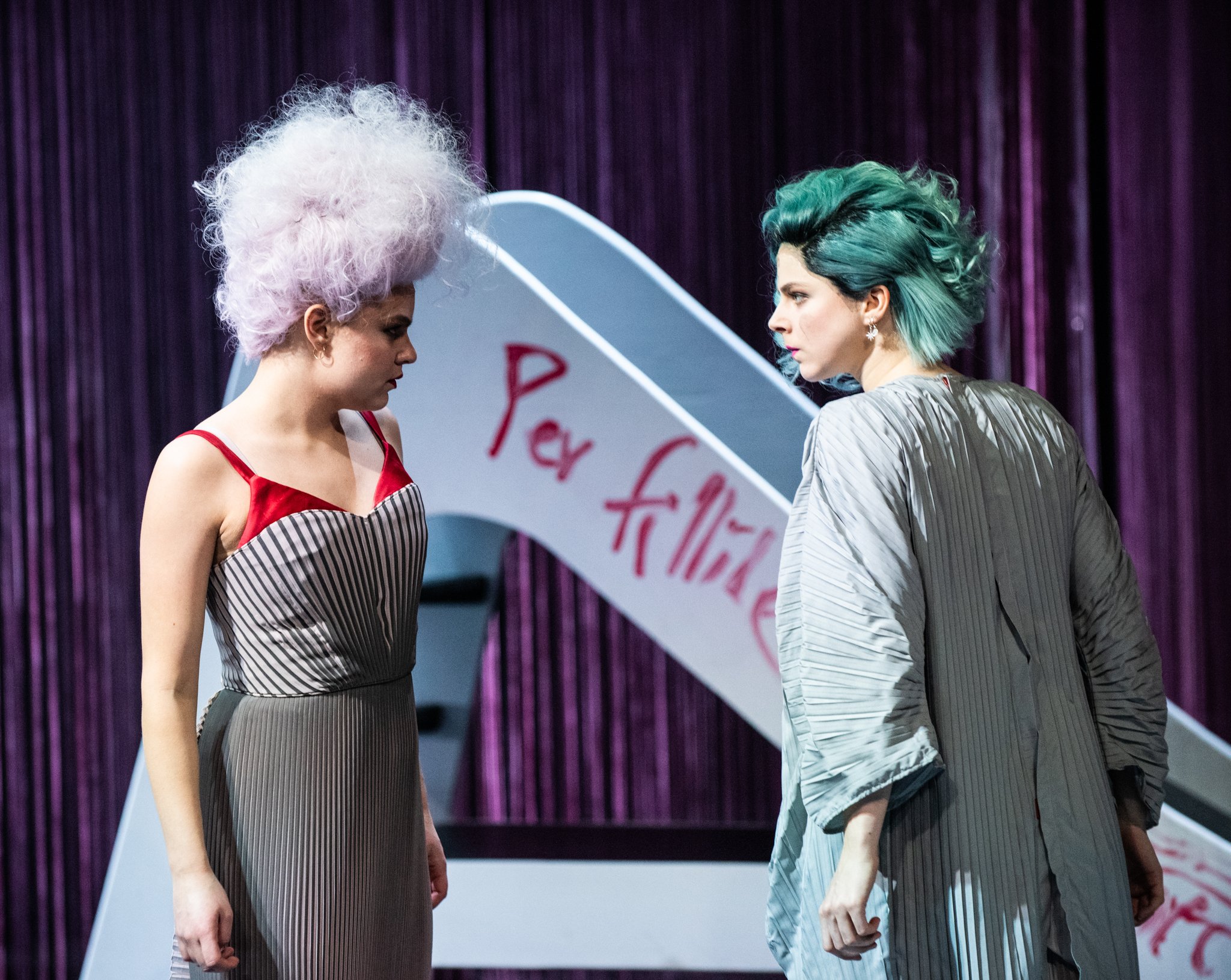 Two women with colorful hair stand close together on a stage with purple curtains, near a white piano with red writing.
