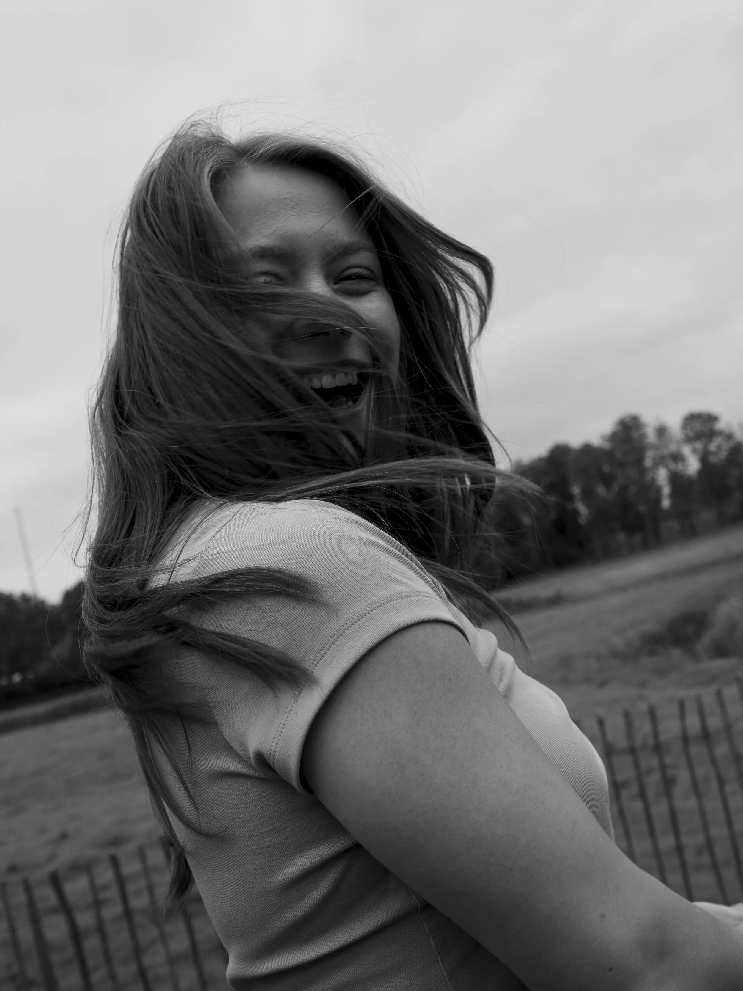 A woman with long hair smiling outdoors in black and white.