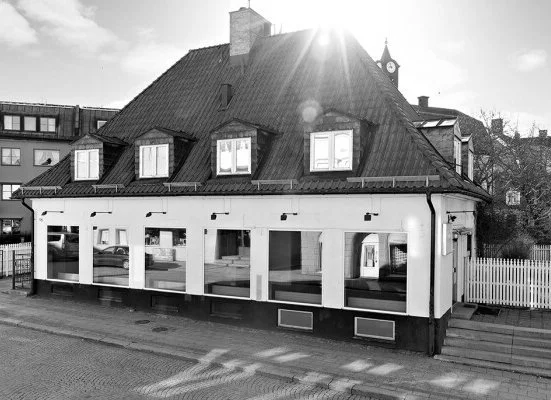 Black and white photo of a two-story house with a steep roof, multiple windows, and a white fence on the right side. The street and sidewalk are visible in front of the house.