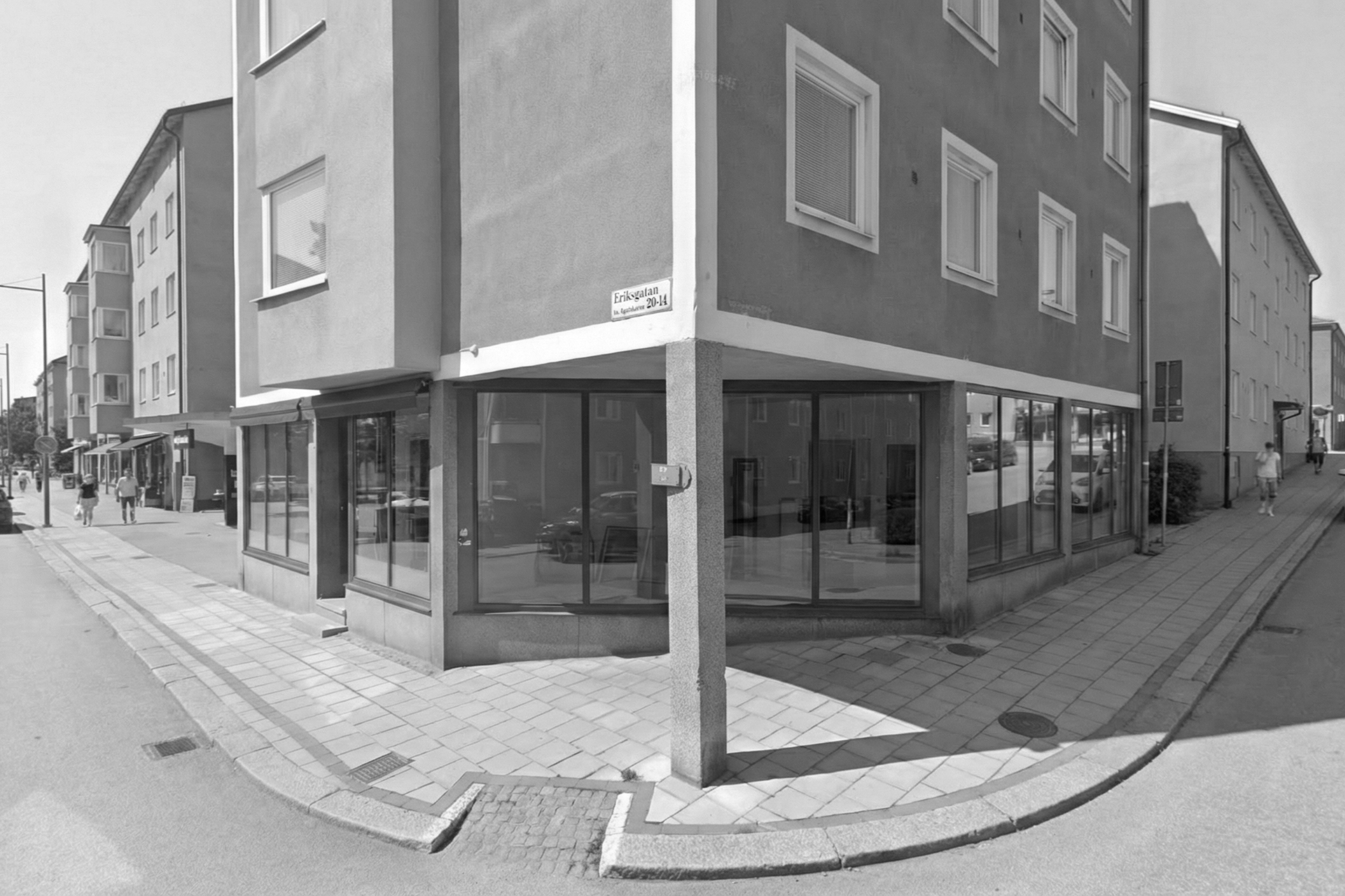 A black and white street view with a corner building featuring large glass windows on the ground floor, residential windows above, and pedestrians walking along the sidewalk.