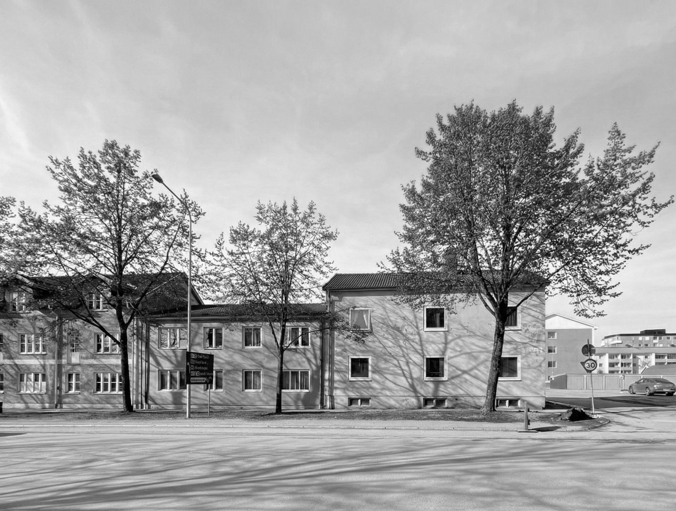 Black and white photo of a residential street with multi-story apartment buildings, trees, traffic signs, a car, and a lamp post.